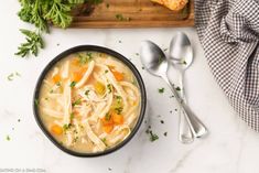 a bowl of chicken noodle soup on a marble counter top with spoons and parsley