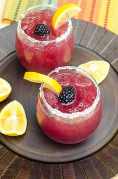 two glasses filled with red drinks and topped with blackberries, lemons and garnishes