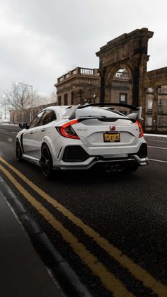 the rear end of a white sports car driving down a road in front of an old building