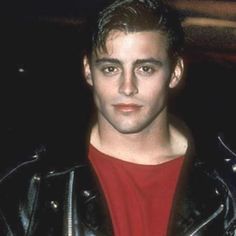 a young man in a leather jacket and red t - shirt looks at the camera