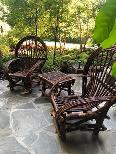 three wicker chairs and a table on a stone patio surrounded by greenery, with trees in the background