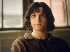 a young man with long hair standing in front of a window and looking at the camera