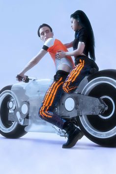 a man and woman riding on the back of a futuristic motorcycle with an orange stripe