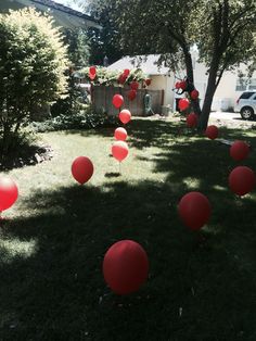 many red balloons are floating in the air near some trees and grass on a lawn