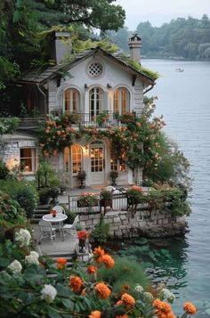 the house is surrounded by flowers and greenery on the water's edge, with an outdoor dining area next to it