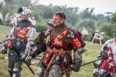 two men dressed in medieval armor and holding swords are walking through the grass with other people behind them