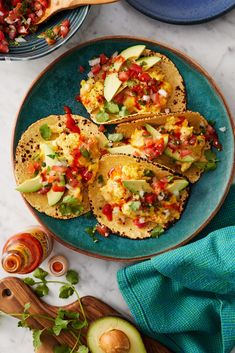 three tacos on a blue plate with avocado and salsa in the background