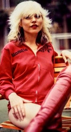 a woman sitting on top of a wooden bench wearing a red jacket and white hair