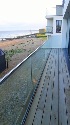 a wooden deck with glass railing next to the ocean and beach side house in the background