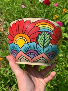 a hand holding up a colorful painted bowl in front of some wildflowers and grass