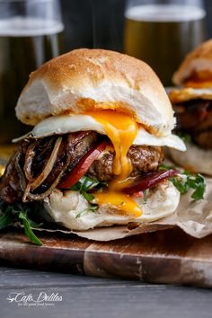 two burgers with meat, cheese and onions on a cutting board next to beer glasses