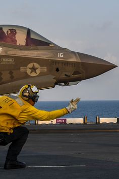 a fighter jet sitting on top of an aircraft carrier next to the ocean with a man in a yellow jacket