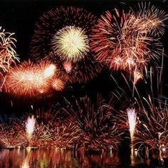 fireworks are lit up in the night sky over water and trees with reflections on it