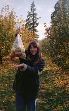 a woman holding a bag full of fruit in an apple orchard with trees behind her