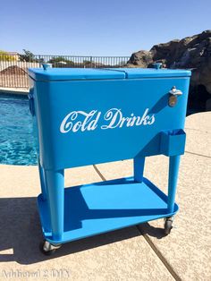 a blue cooler sitting on top of a sidewalk next to a swimming pool with the word cold drinks written on it