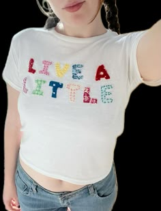 a woman wearing a white shirt with the words live a little written on it in multicolored letters