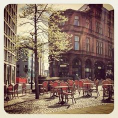 there are many tables and chairs on the sidewalk in front of this building with tall buildings