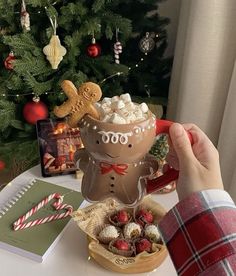 a person holding a mug filled with hot chocolate and marshmallows in front of a christmas tree