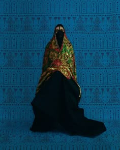 a woman in a black dress with a colorful shawl on her head sitting against a blue wall