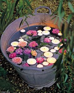 candles floating in a bucket filled with water surrounded by pink and red flowers on the ground