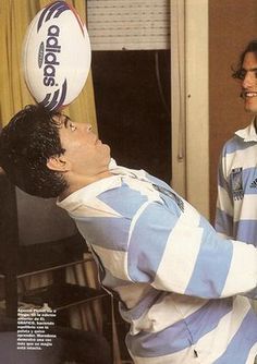 a man balancing a rugby ball on his head while another man watches from the other side