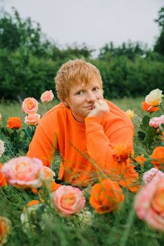 a boy in an orange shirt is sitting in a field of flowers with his hand on his chin