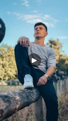 a man sitting on top of a wooden fence next to a tree trunk with his legs crossed