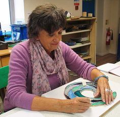 an older woman sitting at a table working on a piece of art