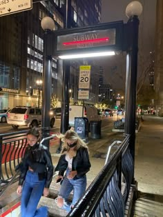 two women are walking down the stairs in front of a subway station sign at night