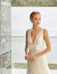 a woman in a white dress standing next to a wall and looking at the camera