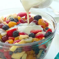 fruit salad with dressing being drizzled over it in a glass bowl on a table