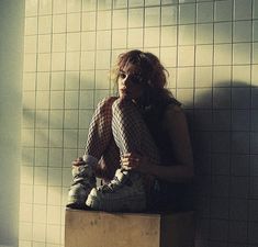 a woman sitting on top of a wooden box in front of a wall with tiled walls