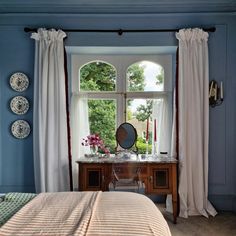 a bedroom with blue walls and white drapes on the windowsills, along with a desk in front of a large window