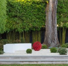 a red sculpture sitting in the middle of a park next to a tree and white bench