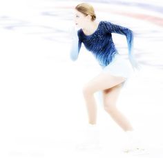 a female figure skating on the ice in a short white skirt and blue sweater with long sleeves