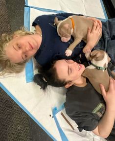 two women laying on the ground with their dogs and one is holding a puppy in her lap