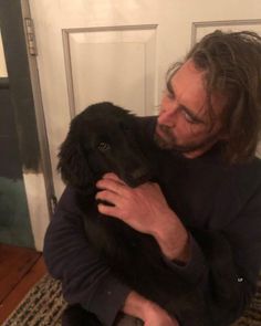 a man sitting on the floor holding a black dog in his arms and looking at the camera