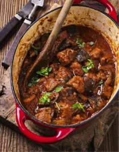 a red pot filled with meat and vegetables on top of a wooden table next to a knife