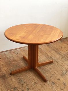 a round wooden table with four legs on a hard wood flooring area next to a white wall