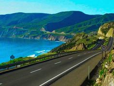 an image of a highway going down the side of a hill by the ocean with mountains in the background