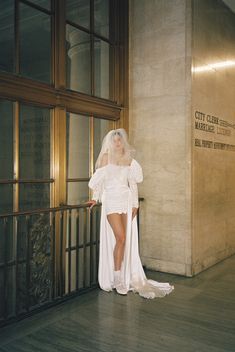 a woman in a white wedding dress standing next to a wall with glass doors and windows