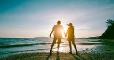 two people standing on the beach holding hands and looking out at the ocean during sunset