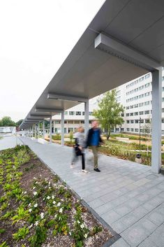 two people walking down a sidewalk near a building with plants growing on the side walk