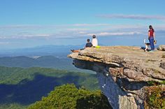 three people sitting on the edge of a cliff looking out at mountains and valleys in the distance