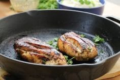 two grilled meats in a skillet on a wooden table next to some vegetables