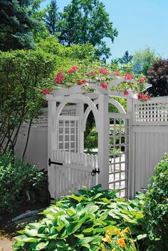 a white garden gate with pink flowers on it and green plants around the fence area