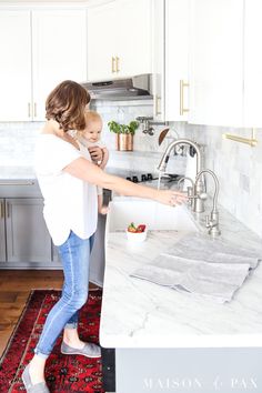 a woman standing in a kitchen next to a sink and counter with a bowl of fruit on it