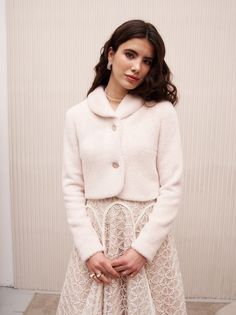 a woman standing in front of a wall wearing a white coat and skirt with buttons