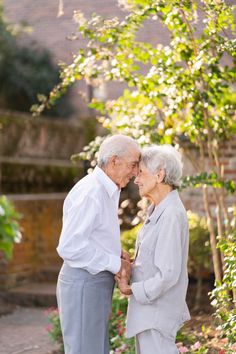 an older couple standing next to each other in front of some trees and flowers on a sunny day