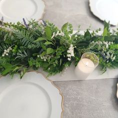 the table is set with white plates and greenery on it, along with a lit candle